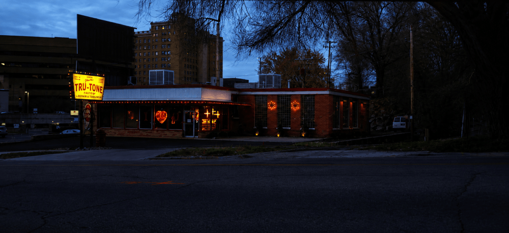 A dimly lit restaurant named Tru-Tone, featuring a neon sign, set against a backdrop of a darkening sky and surrounding buildings.
