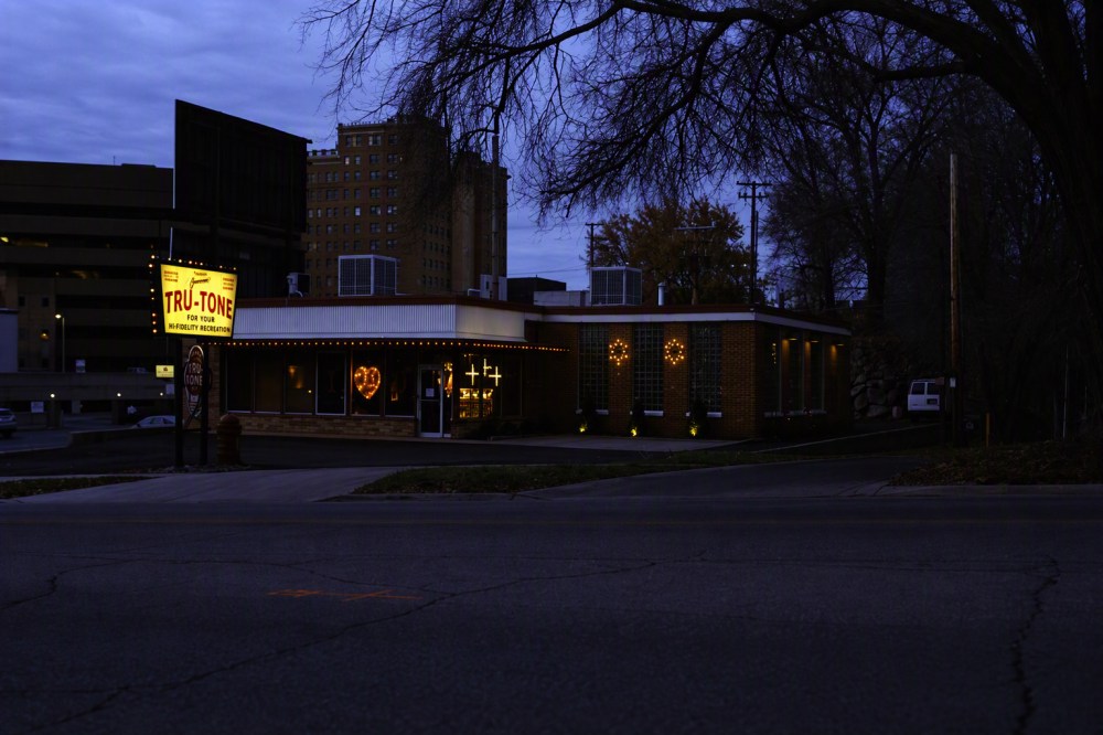 Image of a brick building with illuminated signage reading 'TRU-TONE' at dusk, with a heart-shaped light displayed in the window and a cityscape backdrop.