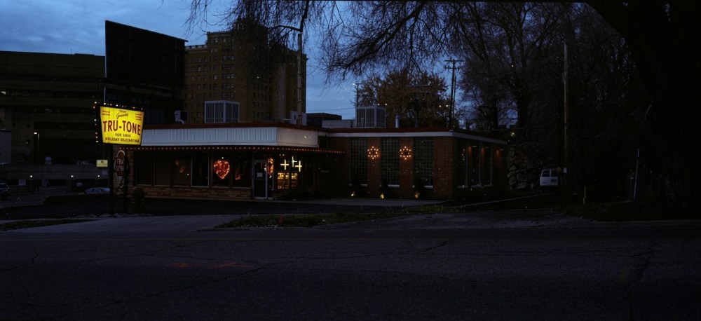 A diner named Tru-Tone illuminated at dusk, featuring a vintage sign and warm exterior lights, surrounded by urban architecture and trees.