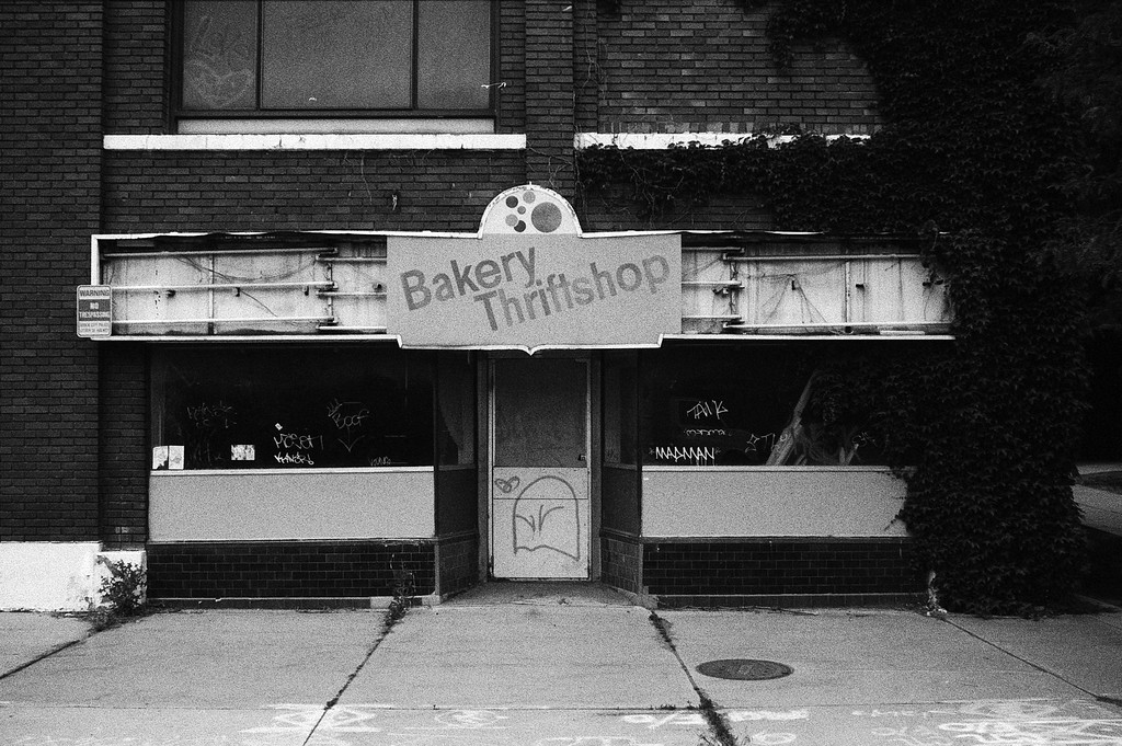 Abandoned thrift shop with a faded 'Bakery Thriftshop' sign, covered in graffiti and surrounded by ivy.
